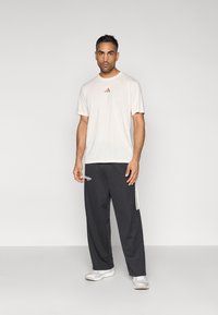 Világos bézs rövid ujjú póló egy kis piros logóval, sötétszürke, széles szárú melegítőnadrággal, fehér oldalsávokkal és logóval párosítva.