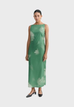 Kobieta o ciemnych włosach, ubrana w zieloną, bezrękawnikową maxi sukienkę z subtelnym wzorem tie-dye, nosząca czarne buty na niskim obcasie na jednolitym tle.