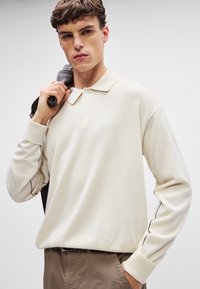Απαλή κρέμ longa-sleeve polo shirt φτιαγμένη από μαλακό ύφασμα με κλασικό γιακά, διαθέτοντας αντίθετες ραφές κατά μήκος των μανικιών.