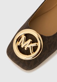 Καφέ επίπεδη υφασμάτινη γόβα με μεγάλο χρυσό κυκλικό έμβλημα, όπου εμφανίζονται με έντονα γράμματα �τα γράμματα "MK" στο μπροστινό μέρος.