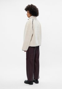 Beige fleece jas met een hoge kraag, contrasterende zwarte stiksels en een losse pasvorm. Gepaard met bordeauxleren wijde broek.