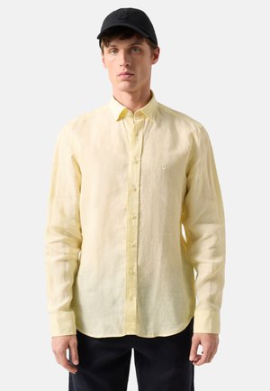 Mand iført en lysegul knap-op langærmet skjorte og sort baseballkasket, står mod en ensfarvet lys baggrund.