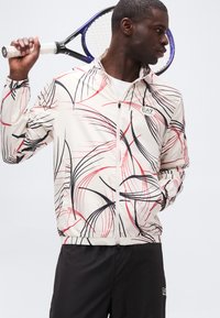 Άνδρας που κρατά ρακέτα τένις στον ώμο, φορόντας λευκό μπουφάν με φερμουάρ και μαύρες και κόκκινες αφηρημένες γραμμές, και μαύρο παντελόνι με τσέπες.