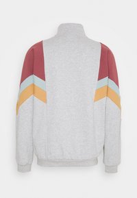 Sudadera gris con cuello alto, que presenta rayas diagonales rojas, azul claro y amarillas en los hombros. Textura suave, puños acanalados.