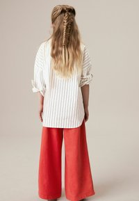 Fehér és fekete csíkos, oversized ing, felgöngyölt ujjakkal, széles piros, rövid nadrággal párosítva, hátulról nézve.