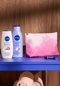 NIVEA shower gel and soft milk bottles in white and light blue, a gradient pink cosmetic bag, and a Labello balm on a blue shelf. Towel below.