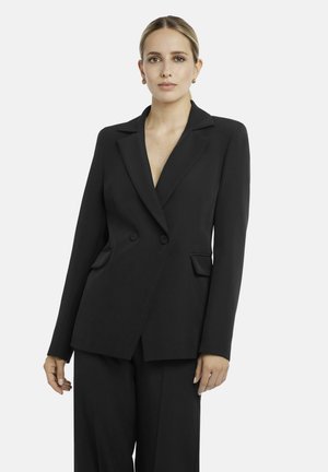 Woman with light hair in a black tailored double-button blazer and matching black trousers stands against a plain light background.