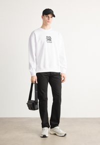Hvit hettegenser med "HUGO"-logo, svarte casual bukser, grå sko og en liten svart crossbody-veske. Modellen står mot en lys bakgrunn.