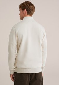 Felpa color beige chiaro con collo alto, polsini e orlo a costine, caratterizzata da una texture liscia e un design semplice, vista da dietro.