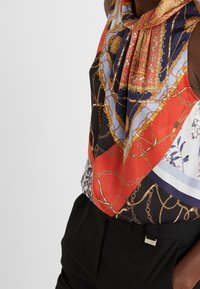 Mouwloze blouse met een gedurfde rode, blauwe en zwarte print met gouden kettingaccenten. Gladde stof met een geplooide halslijn.