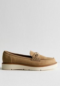 Mocassin en daim beige avec un détail en boucle dorée, semelle blanche plate et silhouette classique. Texture lisse avec des accents de couture minimaux.