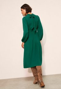 Zelené dlouhé šaty s mašlí na zádech, upnutým pasem a volánkovou sukní. Modelka má na sobě béžové vysoké kozačky.