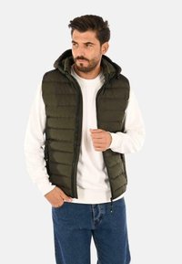 Gilet puffer verde oliva con cappuccio rimovibile, chiusura frontale con zip e tasche laterali, indossato sopra un maglione bianco a collo tondo.