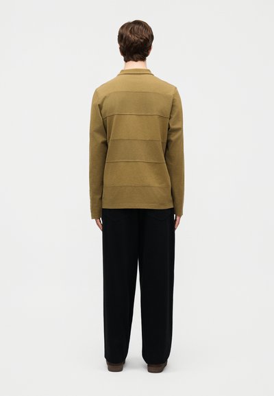 Άτομο που φοράει ελαιδί μακρυμάνικο πουκάμισο και φαρδιά μαύρη παντελόνα, στέκεται με την πλάτη προς τα εμάς μπροστά σε απλό ανοιχτό φόντο.