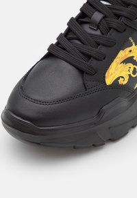 Gros plan d'une basket noire avec des lacets épais et un motif ornemental jaune sur le côté au-dessus de la semelle texturée.