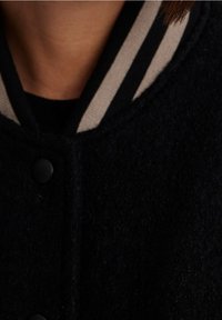 Mustavillainen villatakki, jossa on raidallinen harmaa ja valkoinen kaulus. Siinä on mustat nappikiinnitykset ja pehmeä rakenne.