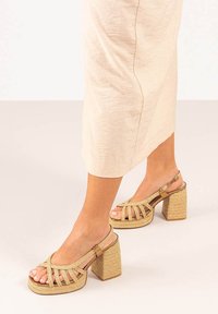 Sandalias de tacón alto de color beige claro con una textura tejida, diseño de punta abierta y un tacón grueso de bloque. Presentan tiras entrelazadas y cierre en el tobillo.