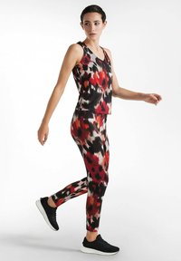 Mujer con top sin mangas de estampado abstracto en rojo, negro y blanco, y leggings, caminando con zapatillas negras sobre un fondo blanco.