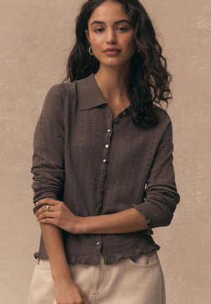 Jeune femme aux cheveux longs et bouclés portant un cardigan en maille marron boutonné et un pantalon beige, debout devant un fond beige uni.