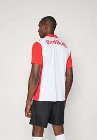 Červený a biely dres s krátkymi rukávmi a logom "Red Bulls" na chrbte, vyrobený z ľahkého materiálu. Skombinovaný s tmavými šortkami a bielymi ponožkami.