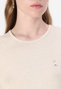 Beige kortermet t-skjorte laget av mykt stoff, med rund hals og en liten sølvdekorasjon på brystet.