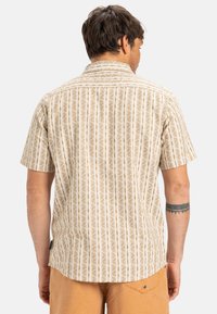 Camisa de manga corta en color crema con un patrón floral vertical en marrón. Presenta un cuello de botón y un corte recto con un ajuste relajado.