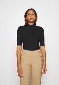 Black ribbed knit top with short sleeves and a mock neck. Worn with beige high-waisted trousers featuring a front button closure.