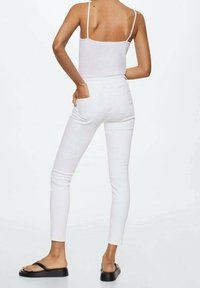 Jeans ajustados blancos con cintura alta y bolsillos traseros estándar, combinados con una camiseta sin mangas blanca. El calzado son chanclas negras con suela plana.