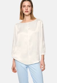 White satin blouse with a loose fit, boat neckline, and 3/4 sleeves. The sleeves feature a gathered detail at the cuffs. Paired with light blue trousers.