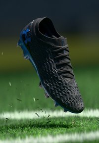 Scarpa da calcio atletica nera con tomaia texture, accenti blu chiaro e suola stampata, rappresentata in volo con detriti d'erba.