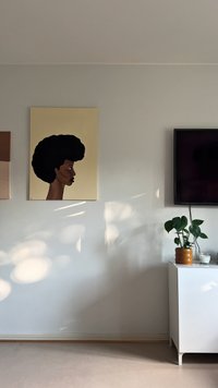 Platno z umetniškim delom, ki prikazuje profil ženske z črnim kodrastim afrom na svetlo rumeni podlagi. Moderna notranjost z belim omarico in rastlino.