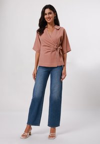 Top avvolgente color rosa con vita legata, maniche ampie e cuciture a contrasto, abbinato a jeans a vita alta blu svasati e sandali bianchi con tacco a blocco.