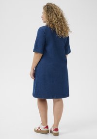Denim dress in dark blue, knee-length, short sleeves, straight cut, and visible stitching. Worn with beige platform sneakers with red accents.