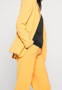 Gul skräddarsydd kavaj och matchande byxor i ett mjukt tyg, bärs över en svart topp. Outfiten har rena linjer och en strukturerad design.