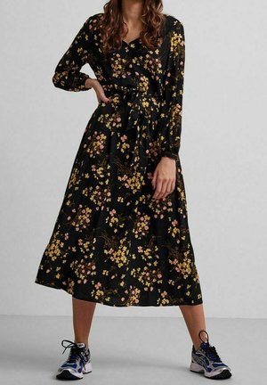 Vrouw die een zwarte midi-jurk met lange mouwen draagt, bedrukt met gele en roze bloemen, gecombineerd met blauwe en zilveren sportschoenen.