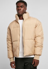Beige puffjakke med høy krage, glidelås og horisontal quilted mønster. Har elastiske mansjetter og en lett cropped passform.