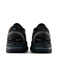 Μαύρα αθλητικά παπούτσια με γκρι λεπτομέρειες, διαθέτουν μαλακή σόλα και υφασμάτινη υφή. Η φτέρνα φέρει την ένδειξη "1000" και ένα λογότυπο σε μπλε χρώμα.