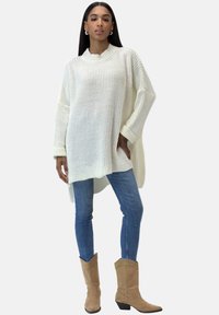 Maglione bianco oversize lavorato a maglia con colletto e polsini a coste, abbinato a jeans skinny blu e stivali da cowboy color cammello a metà polpaccio.