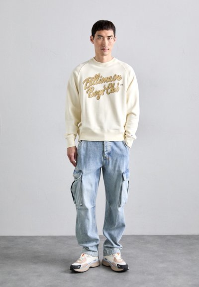 Man wearing cream sweatshirt with "Billionaire Boys Club" text, light blue cargo jeans, and white sneakers standing against a plain gray wall.