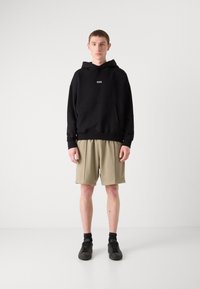 Black hooded sweatshirt with front pocket, paired with beige knee-length shorts. Model wearing black shoes with dark socks, standing on a light background.