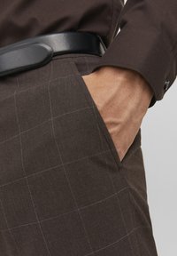 Pantaloni a quadri marroni con una texture liscia, tasche laterali e una cintura in pelle nera. Il design presenta sottili linee a griglia bianche.