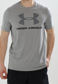 Camiseta de manga corta gris con una textura suave, que presenta un gran logo negro de Under Armour en el pecho. Diseño clásico de cuello redondo.