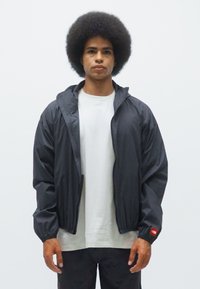 Veste légère noire avec capuche, poignets élastiques et accent rouge sur le poignet gauche, portée sur un t-shirt blanc uni et un pantalon noir.