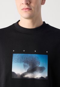 Felpa nera con una grafica frontale che presenta un motivo vorticoso in diverse tonalità di blu e nero, con il testo "HUGO" sopra l'immagine.