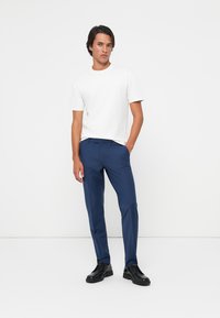 Blaue maßgeschneiderte Hosen aus glattem Stoff, kombiniert mit einem weißen T-Shirt mit Rundhalsausschnitt. Die Schuhe sind schwarz und haben ein Schnürdesign.