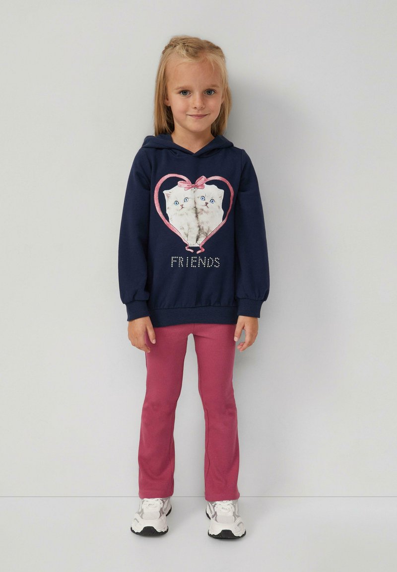Jong meisje met lang blond haar, gekleed in een marineblauwe hoodie met een kattenprint en de tekst "Friends", roze broek en witte sneakers, staande tegen een witte muur.