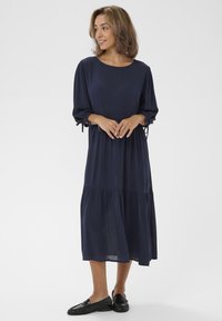 Marineblaues, knielanges Kleid mit Dreiviertelärmeln und gerafftem Detail an der Taille. Glatter Stoff, lässiger Schnitt, mit seitlichen Bindungen. Schwarze Slipper.
