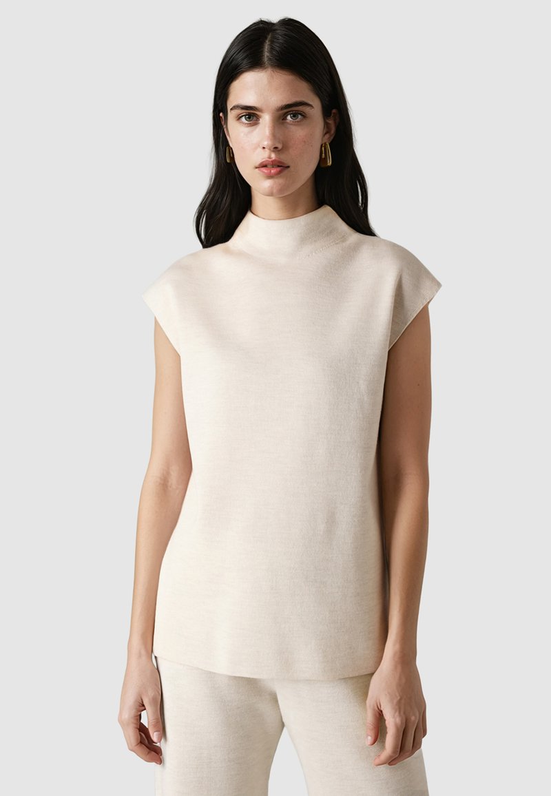 Femme aux cheveux foncés portant un haut beige sans manches à col montant et un pantalon assorti, debout devant un fond gris clair.