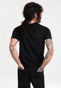 Camiseta negra de manga corta hecha de algodón con escote redondo. La persona tiene tatuajes en los brazos y las manos en los bolsillos, de espaldas a la cámara.