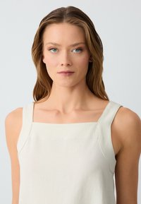 Giovane donna con capelli castano chiaro che indossa un top senza maniche beige, che guarda dritto davanti a sé con un'espressione neutra, sfondo grigio chiaro.